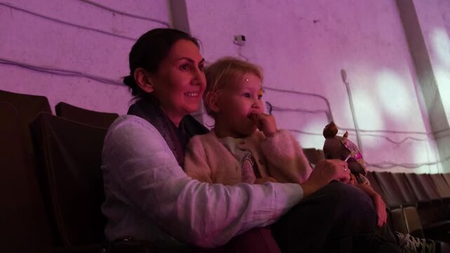 Candid moment of a woman with her young daughter on her lap enjoying a live show in a dark concert hall with colorful stage lighting, smiling and reacting to the performance
