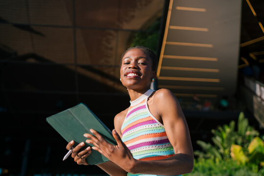 Smiling African woman using tablet outdoors in sunlight, expressing digital creativity, confidence, and remote work freedom with modern tech tools and vibrant personality. - Powered by Adobe