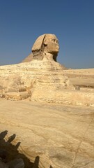 A large statue of the Great Sphinx stands on sandy ground. Pyramids are visible behind it. Many visitors look at the statue. The weather is sunny and clear with no clouds in the sky