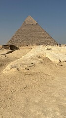 People explore the area around the Great Pyramid of Giza, one of the oldest and largest pyramids in Egypt. The sun shines brightly in the sky as the sand covers the ground