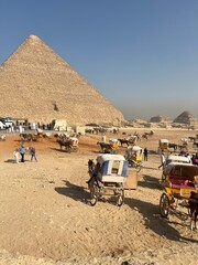 Visitors spend time near the pyramids in Egypt while horses and carriages are ready for rides. People walk around and enjoy the warm weather at this famous historical site