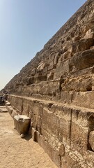 Visitors walk near the base of the Great Pyramid of Giza. The ancient structure stands tall with large stone blocks visible. Sunlight shines brightly on the scene, showing its historic details