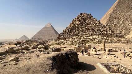Visitors walk around the pyramids of Giza, observing ancient structures and stones. The scene shows the desert landscape with clear skies and distant pyramids. People interact with the site