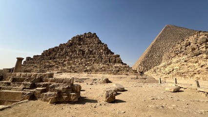 Visitors walk among the stone structures of the pyramids in Giza. The sun is high and the ground is dry. Some stones are scattered, and the pyramids stand tall against the clear sky
