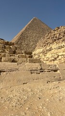 Tourists walk near ancient stone structures while viewing the Great Pyramid of Giza standing tall in the background on a sunny day. The scene shows historic landmarks and desert landscape