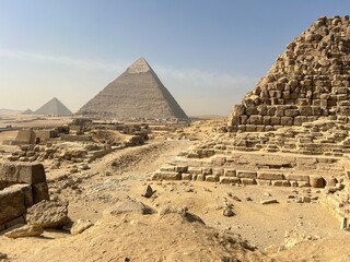 Visitors walk among the ancient stone structures at Giza. The iconic pyramids stand tall in the distance under a blue sky. Sand covers the ground and hints of ancient history surround the area