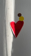 A heart made of red paper sits on a wall, surrounded by circular shapes in brown and yellow. Sunlight casts shadows, highlighting the vibrant colors in a simple space