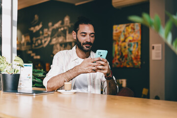 Laughing man with beard holding smartphone in café, coffee on counter. Conceptual image of digital joy, independence, individuality, and authentic modern communication lifestyle.
