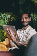Man leans back, smiling at tablet screen with natural charm, surrounded by lush green wall, symbolizing harmonious connection between tech, comfort, and joyful living. © BullRun
