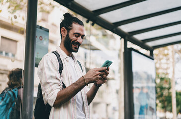 Freelancer reads smartphone with smile, standing in sunlight under bus stop roof, symbolizing effortless connection, positivity and tech-enabled daily mobility.