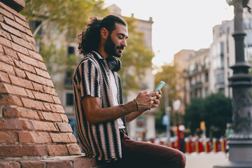 Fototapeta premium Smiling man chats on phone while seated outside, enjoying digital conversation, relaxed moment and mobile connection in casual city environment.