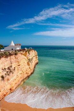 A breathtaking aerial view captures the iconic white-washed Church of Nossa Senhora da Rocha perched dramatically atop golden sandstone cliffs in the Algarve, Portugal.
