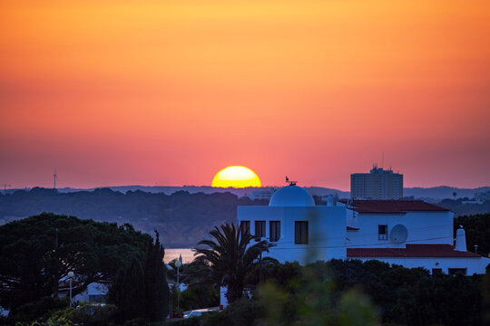 A stunning sunset over Ferragudo in the Algarve, Portugal, where the sun dips below the distant hills, painting the sky in vibrant hues of orange and pink.
