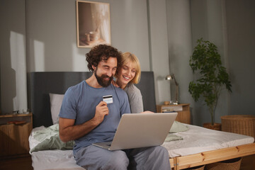 Two people are shopping online in their bedroom, smiling and laughing, while one holds a credit card and the other uses a laptop. They are relaxed and enjoying their time together.
