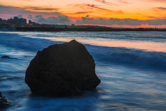A breathtaking sunset view from Praia Pintadinho in the Algarve, Portugal, where a solitary, dark rock stands silhouetted against the vibrant orange and pink sky.