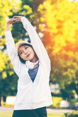 Asian woman wearing wireless headphones, smiling while relaxing in a park after workout and stretching, enjoying music and peaceful outdoor moment of contentment and well-being. 