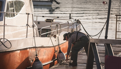 Senior man wearing overalls fastening rope to boat cleat on marina dock with fenders, copy space