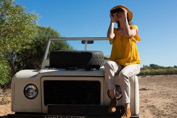 Woman wearing straw hat holding binoculars on vehicle hood with spare tire in scrubland, copy space