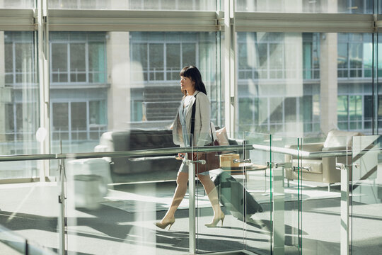 Solo businesswoman walking along glass-railed airport walkway rolling suitcase past sofas, windows