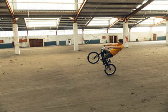 Male rider performing wheelie on BMX bicycle in warehouse wearing yellow T-shirt, copy space
