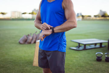 Mid-thirties man adjusting smartwatch on wrist on grassy field with kettlebells, sandbags, plyo box