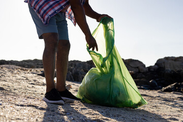 Senior African American man in checked shirt bending over at beach collecting trash with green bag