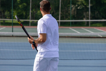 Mid adult male standing near net on tennis court with racket by chainlink fence wearing smartwatch