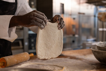 African American male chef holding flat dough above floured countertop with mixing bowl in kitchen