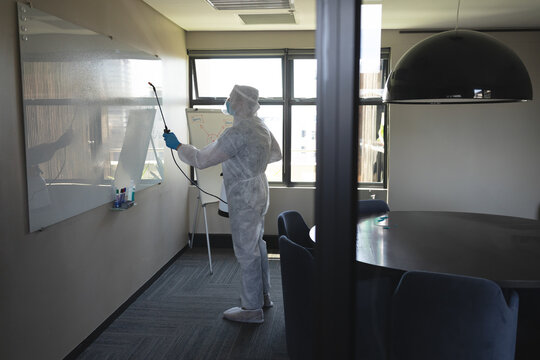 Man wearing protective suit spraying disinfectant on whiteboard using spray wand in conference room
