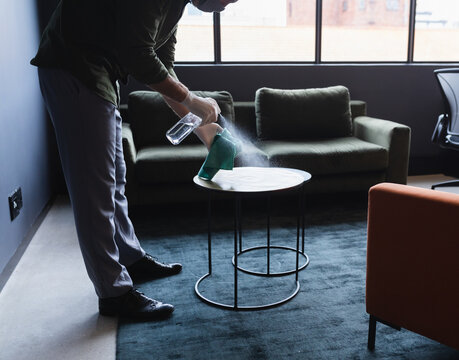 Man wearing gloves cleaning side table in office lounge with spray bottle and cloth, copy space