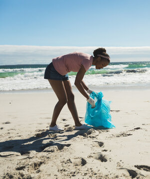 African American female with gloves bending over on beach picking up litter into blue trash bag
