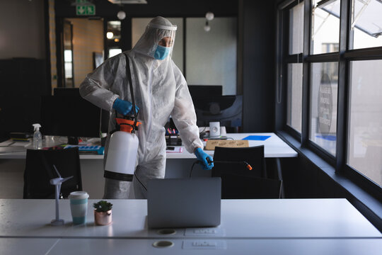 Male cleaning technician in protective suit sanitizing office with sprayer at laptops and monitors - Powered by Adobe