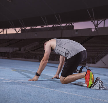 Male athlete crouching at starting blocks on blue track in stadium with running blade, sports watch