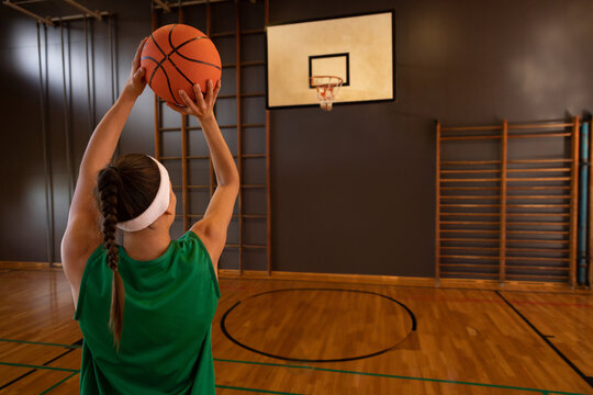 Female athlete shooting basketball toward hoop wearing green jersey on gym court with wall bars