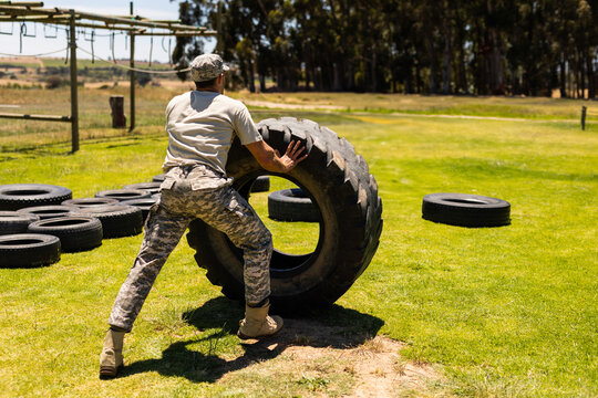Male trainee pushing large tractor tire across training field near tires wearing combat boots