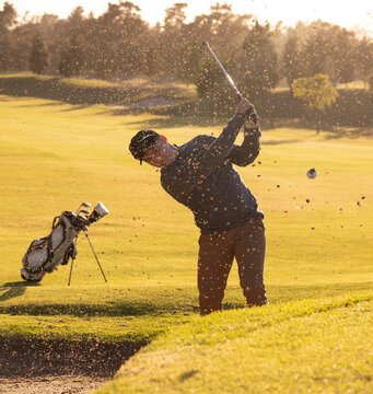 Male golfer swinging club, hitting golf ball from sand bunker on golf course near golf bag