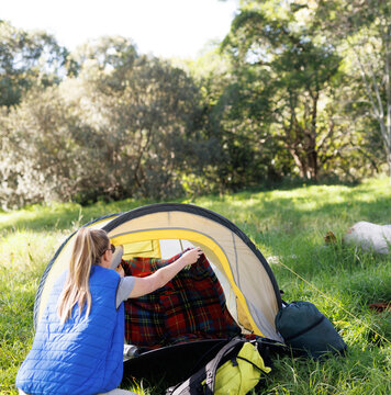 Woman crouching in grassy clearing at dome tent entrance pulling plaid blanket inside, copy space