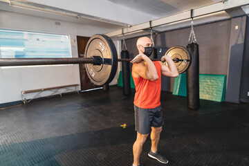 Adult male wearing red tank top holding barbell loaded 15kg plates in gym with punching bags