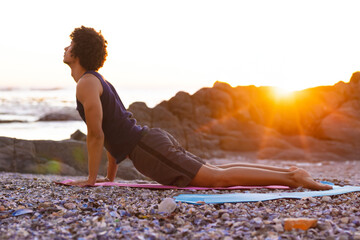 African American man in sleeveless top arching on yoga mats on pebble beach with ocean waves