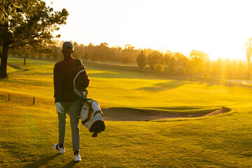 African American man in golf attire walking across fairway at sunrise carrying golf bag on shoulder