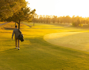 Adult man walking across golf course at sunrise wearing pullover, carrying golf bag, copy space