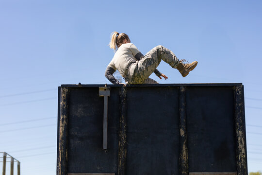 Adult female military trainee vaulting over wooden obstacle wall with brackets on training ground