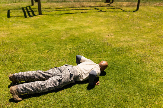 African American man wearing military uniform performing push-up on grassy field by wooden obstacle