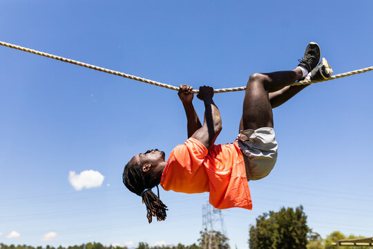 African American man in orange shirt hanging inverted from thick rope on outdoor obstacle course