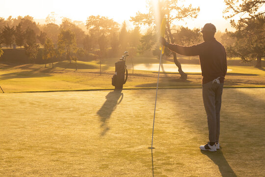African American man gripping flagstick on putting green with golf bag near pond, copy space