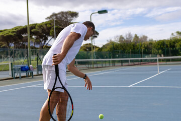 Mid-adult man in tennis outfit bouncing ball with racket on blue hardcourt by fence, copy space