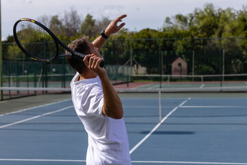 Mid adult man serving tennis ball on tennis court by net, holding racket, wearing smartwatch