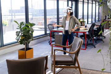 Asian woman standing in office wearing virtual reality headset and gesturing near wooden armchairs
