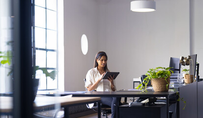 Asian woman holding tablet and reading at modern office desk with monitor keyboard and plants