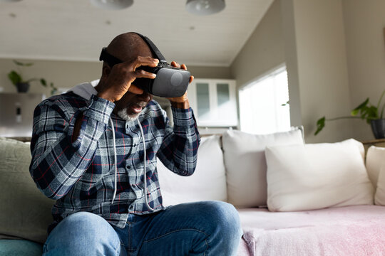Senior African American man lifting VR headset while sitting on sofa beside kitchen cabinets